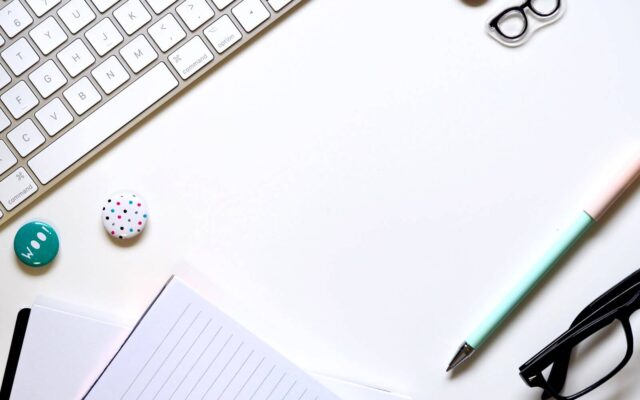 Bild eines weissen Schreibtisches von oben, in der oberen linken Ecke eine Tastatur, unten ein Block, ein Stift und eine Brile, in der mitte zwei farbige Buttons.